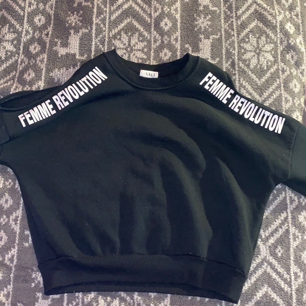 A black sweater with the shoulder open and the shirt say “Femme Revolution”